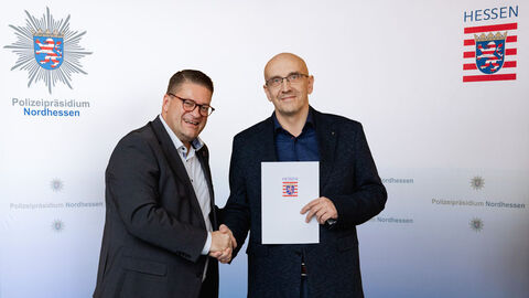 Abteilung Einsatz des Polizeipräsidiums Nordhessen mit neuer Leitung: Rainer Neusüß folgt auf Uwe Papenfuß; Auf dem Foto zu sehen sind der nordhessische Polizeipräsident Marco Bärtl  und der neue Leiter der Abteilung Einsatz, Rainer Neusüß, mit einer Urkunde in der Hand, stehend vor einer weißen Wand mit den Logo des Polizeipräsidiums Nordhessen und dem Hessen-Wappen