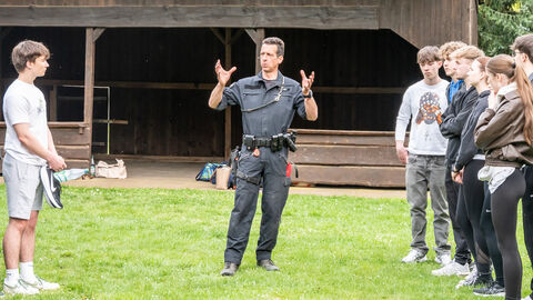 Teilnehme der CopChallenge stehen rechts und link auf einer grünen Wiese während ein uniformierter Diensthundführer Auskünfte aus dem Polizeialltag teilt. Im Hintergrund sieht man eine dunkle Holzhütte. 