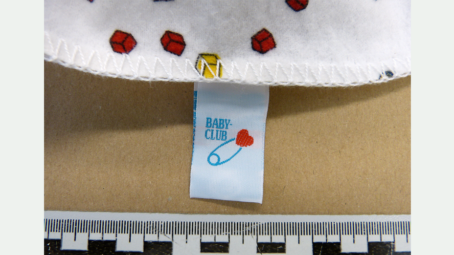 Ein kleines Stück einer weißen Babydecke mit roten und gelben Würfeln darauf und das Etikett mit der Abbildung einer blauen Sicherheitsnadel mit rotem Herz, schräg darüber der Schriftzug BABYCLUB