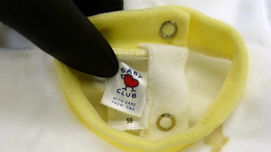 Eine schwarz behandschuhte Hand hält das Etikett im Halsausschnitt eines Baby-Stramplers, auf dem Etikett ist ein rotes Küken mit weißem Schnabel, darüber der Schriftzug BABY, darunter der Schriftzug CLUB, WITH CARE FROM C&A und die Größe 56