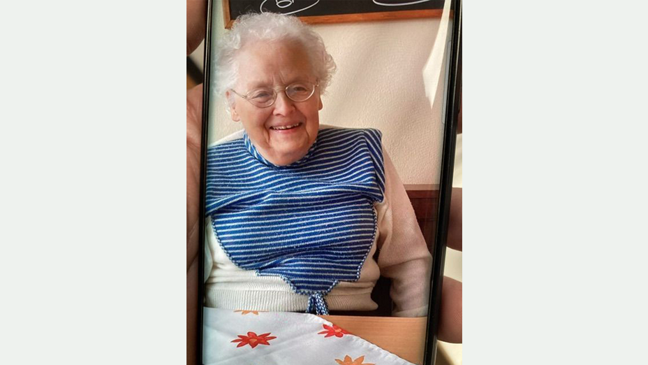 87-jährige Frau mit kurzer grauer Dauerwelle und Brille in einem beigen Pullover mit blau-weiß gestreiften Einsatz