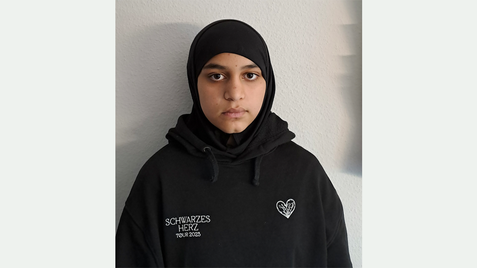 Eine 13-Jährige mit dunklen Augen, schwarzem Kopftuch und schwarzem Kapuzenpullover mit der weißen Aufschrift SCHWARZES HERZ TOUR 2023 und weißes eingesticktes Herz